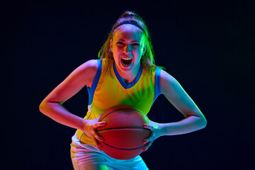 Concentrated and motivated young girl, basketball player in uniform posing with ball against black...