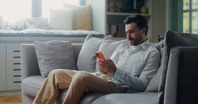 A stylish bearded man sits on the sofa at home and starts typing on his mobile phone, smiling and enjoying his leisure time