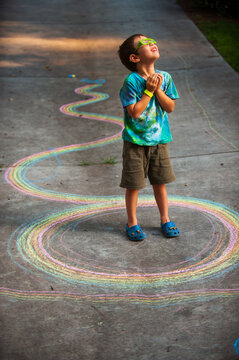 A boy wearing a tie dyed shirt stares at the sky with 3-D glasses while standing inside a design drawn with multicolored chalk on a driveway