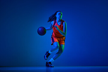 Dynamic image of young girl, basketball player in uniform in motion, playing over blue studio background in neon light. Concept of professional sport, action and motion, game, competition, hobby, ad