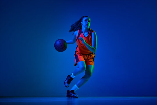 Dynamic image of young girl, basketball player in uniform in motion, playing over blue studio background in neon light. Concept of professional sport, action and motion, game, competition, hobby, ad