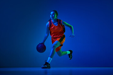 Concentrated female basketball athlete training, dribbling ball against blue studio background in...