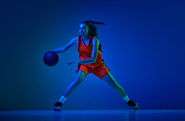 Concentrated female basketball athlete training, dribbling ball against blue studio background in...