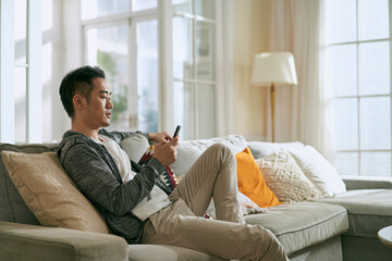 young asian man sitting on family couch at home using mobile phone