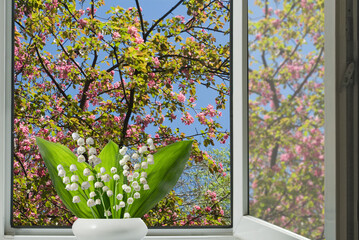  beautiful white flowers of lilies of the valley against the background of an open window behind which a flowering tree can be seen