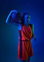 Portrait of young girl, basketball player in uniform posing with ball against blue studio...