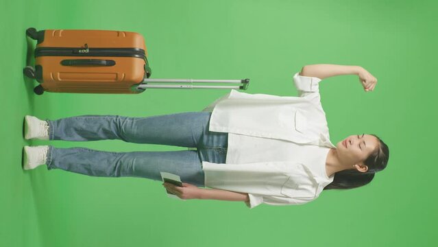 Full Body Of Asian Female Traveler With Luggage And Passport Smiling And Flexing Her Bicep While Standing In The Green Screen Background Studio
