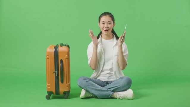 Full Body Of Asian Female Traveler With Luggage And Passport Smiling And Saying Wow While Sitting In The Green Screen Background Studio
