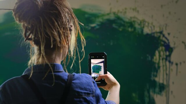 A talented red-haired female artist with dreadlocks takes a photo of a finished masterpiece on a smartphone in a loft studio. Creating an abstract painting for the Internet and an online exhibition.