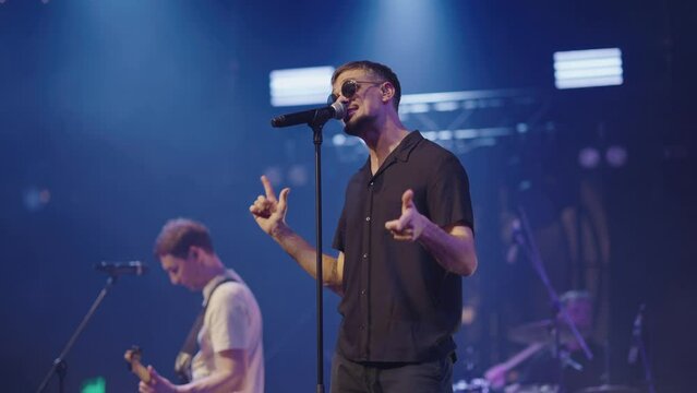 Attractive lead singer of band in dark glasses and black shirt performs on stage of nightclub. Bright spotlights. Slow motion