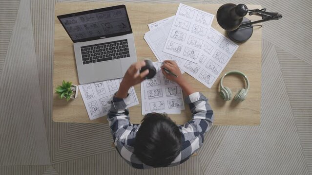 Top View Of Asian Male Artist Drinking Coffee While Drawing Storyboard For The Film On The Table With A Laptop And Headphones In The Studio
