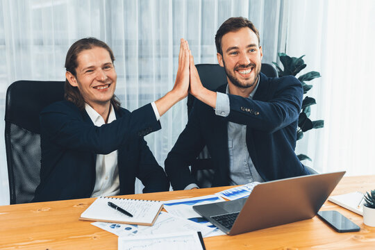Businessmen celebrate success with joyous expressions, celebratory high-five hand gesture in office after analyzing data or achieving target. Teamwork and planning lead to successful outcome. Entity