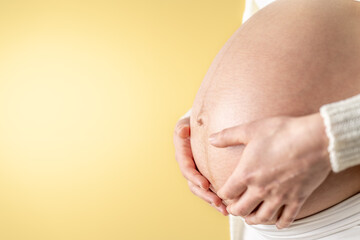 Pregnant woman gently holding her naked round belly with her hands in last month of pregnancy. Third trimester - week 36. Close up. Side view. Yellow background. Bright shot.