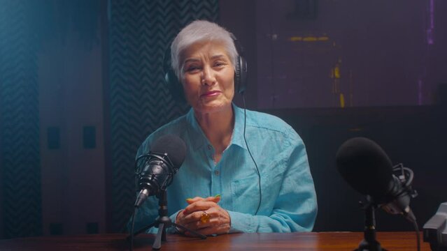 Portrait: Serious old mature blogger woman in wireless headphones recordings podcast in a professional recording studio. An elderly lady on a radio interview. DJ creates live streaming on the Internet