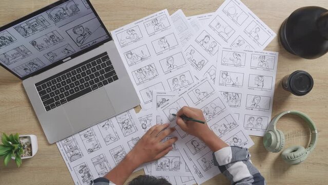 Close Up Top View Of Asian Male Artist Drawing Storyboard For The Film On The Table With A Laptop And Headphones In The Studio
