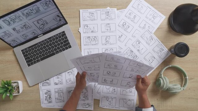 Close Up Top View Of Male Artist'S Hands Holding Sketching Papers Choosing The Best Storyboard For His Project On The Table With A Laptop And Headphones In The Studio
