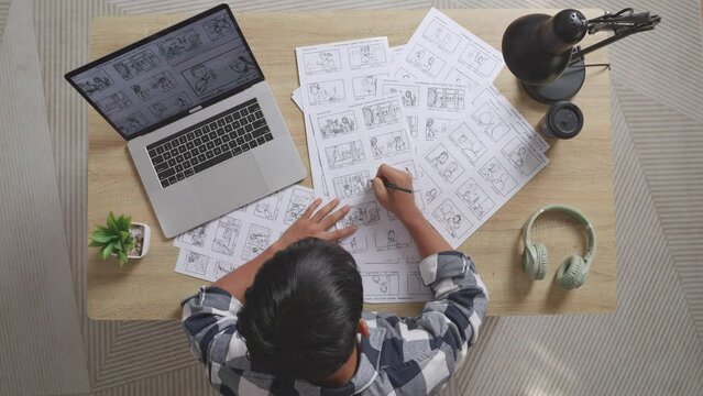 Top View Zoom In Shot Of Asian Male Artist Drawing Storyboard For The Film On The Table With A Laptop And Headphones In The Studio
