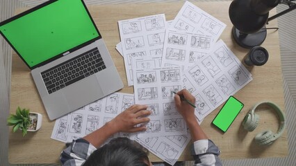 Top View Zoom Out Shot Of Asian Male Artist Drawing Storyboard For The Film On The Table With Green Screen Smartphone And Laptop In The Studio
