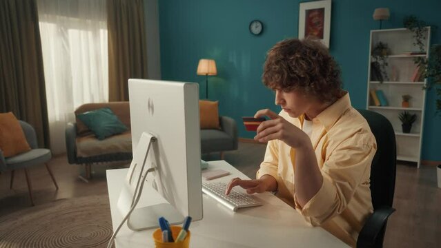 A young guy uses a computer and a credit card to pay for online purchases. The man makes a victory gesture, the payment was successful. No need to leave home to go shopping. Slow motion.