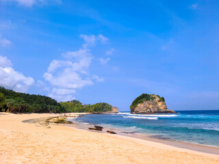 View of beautiful beach with white sand, rocks, green foliages and calm waves under clear blue sky. Summer vacation concept. Ngudel Beach, Malang, Indonesia