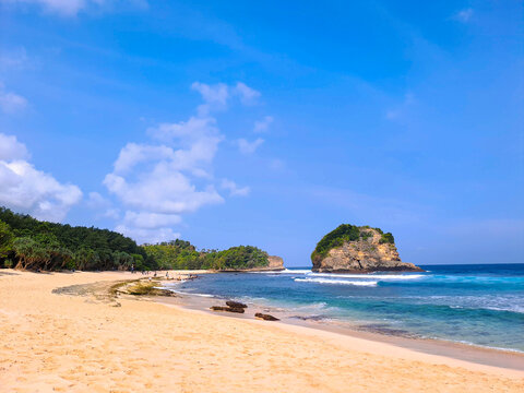 View of beautiful beach with white sand, rocks, green foliages and calm waves under clear blue sky. Summer vacation concept. Ngudel Beach, Malang, Indonesia