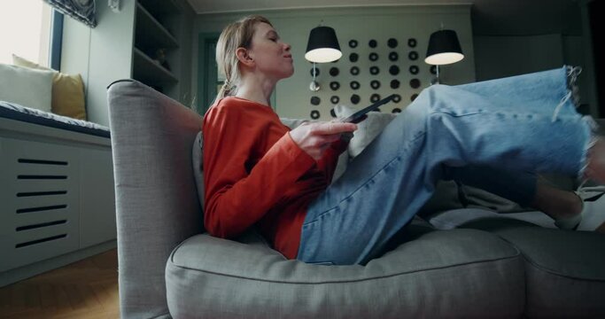 A young woman in casual clothes sits on the sofa and smiles while opening her laptop and starting typing. The video moves from a close-up of women's legs to a general plan