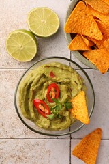 Bowl of delicious guacamole with chili pepper, nachos chips and lime on white tiled table, flat lay