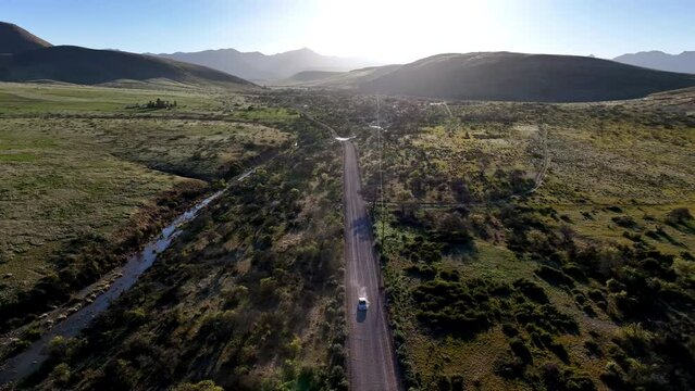 Aerial shot of car driving on dirt road in Willcox, Arizona, wide drone shot with mountains in the background and dust behind the car into the sun