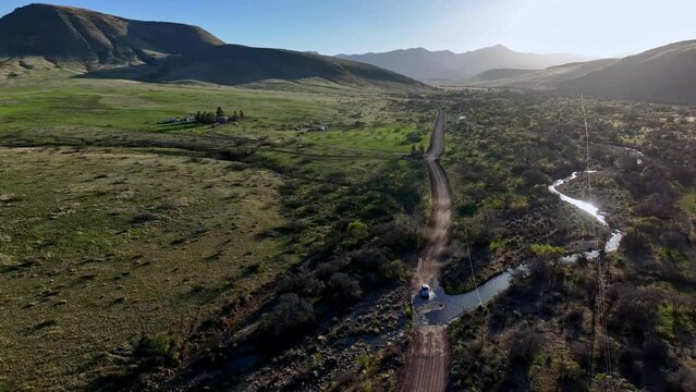 Aerial shot of car driving towards camera on dirt road in Willcox, Arizona, wide rotating drone shot with mountains in the background and dust behind the car
