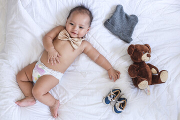 Top view of cute little baby lying on bed wearing bow tie and diaper with knitted toy bear, shoes...