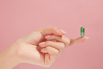On light pink background, a green capsule standing on a finger of hand model. Medicine and health...