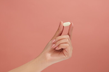 Closeup view of female hand model holding a white pill against pink background. Blank space for...