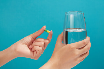 Minimal scene of a yellow-green capsule is held by a hand model, a glass filled with water in another hand. Vitamin can increase quality of life