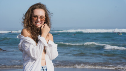Portrait of a wealthy young woman in a white shirt and blue bikini, standing on the Bali coast of the ocean on a sunny day against the blue sky, smiling and looking. Traveling in the southern Asia.