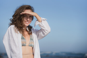 Portrait of a wealthy young woman in a white shirt and blue bikini, standing on the Bali coast of the ocean on a sunny day against the blue sky, smiling and looking. Traveling in the southern Asia.