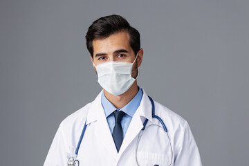 A male doctor in a white coat and medical mask and sterile gloves looks at the camera on a gray isolated background, copy space, space for text
