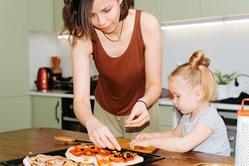 Young mother and daughter preparing pizzas together in kitchen, little caucasian girl and mother adding toppings to dough. Family cooking hobby