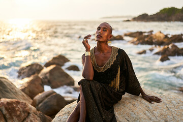 Androgynous ethnic fashion model in luxury dress, jewelry sits on rocks by ocean. Gay black person...