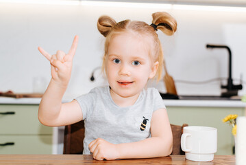 Funny child girl showing rock goat gesture while sitting at table in kitchen and looking at camera