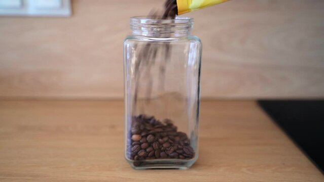 Freshly roasted coffee beans pouring into a jar from a package in the kitchen. A wide angle shot in front of the jar. Coffee beans falling into the container.