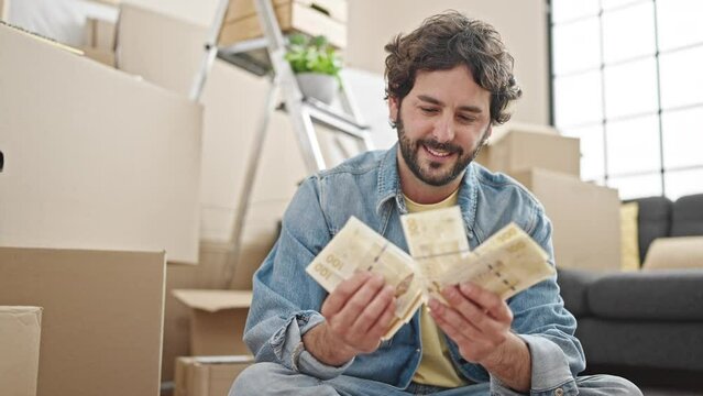 Young hispanic man counting denmark krone banknotes sitting on floor at new home
