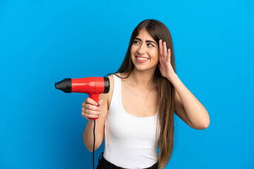 Young caucasian woman holding a hairdryer isolated on blue background listening to something by putting hand on the ear