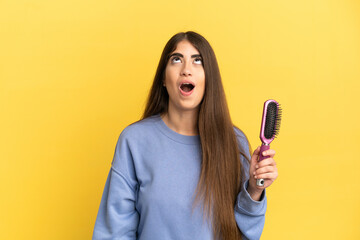 Young caucasian woman holding hairbrush isolated on blue background looking up and with surprised expression - obrazy, fototapety, plakaty