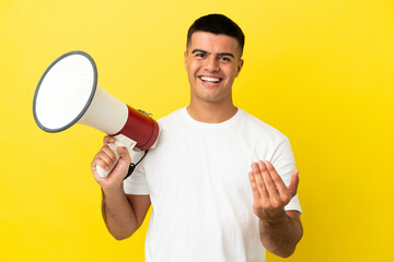 Young handsome man over isolated yellow background holding a megaphone and inviting to come with...