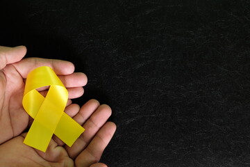 Human hands holding yellow ribbon color on dark black background. Bone cancer awareness and suicide prevention hope concept. - obrazy, fototapety, plakaty