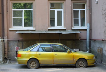 An old yellow car with deflated wheels is parked against the wall of a residential building, Tallinn Street, Saint Petersburg, Russia, June 14, 2023