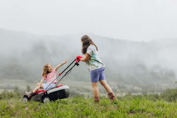 A woman in boots with her child in the form of a game mows the grass with a lawnmower in the garden against the background of mountains and fog, garden tools concept