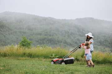 A woman in boots with her child in the form of a game mows the grass with a lawnmower in the garden against the background of mountains and fog, garden tools concept