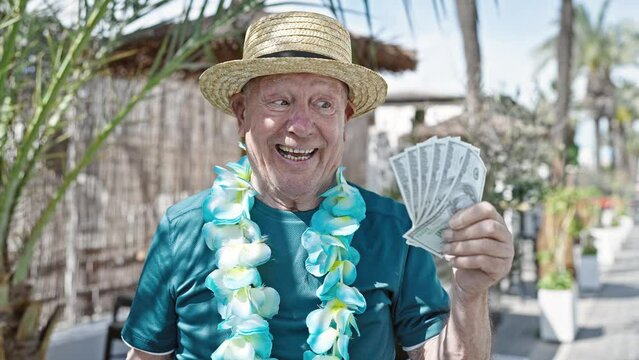 Senior grey-haired man tourist wearing summer hat and hawaiian lei holding dollars at street
