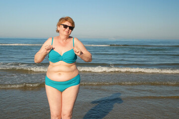 Plus size senior woman in swimsuit dancing on the sea beach in sunny day. Happy people in vacation...